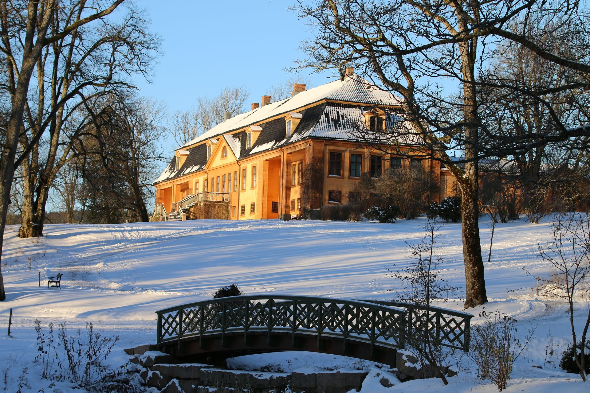 Snow-covered manor house with warm orange walls and a small bridge in the foreground under bare trees.