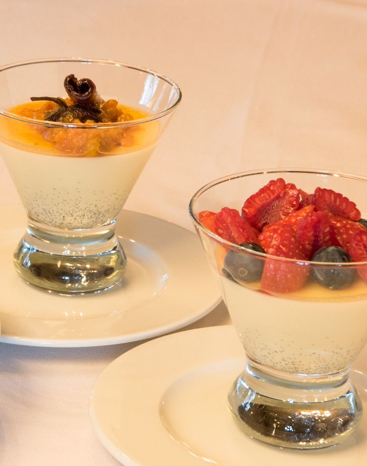 To dessertkopper i glass står på hvite tallerkener, en toppet med fruktkompott og en med friske bær på et hvitt bord.