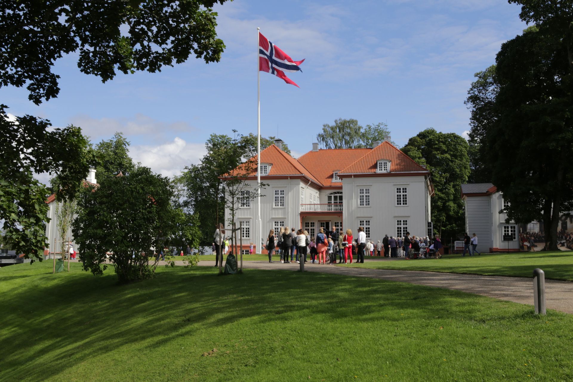 Et norsk flagg vaier over en hvit bygning; folk samles på plenen.
