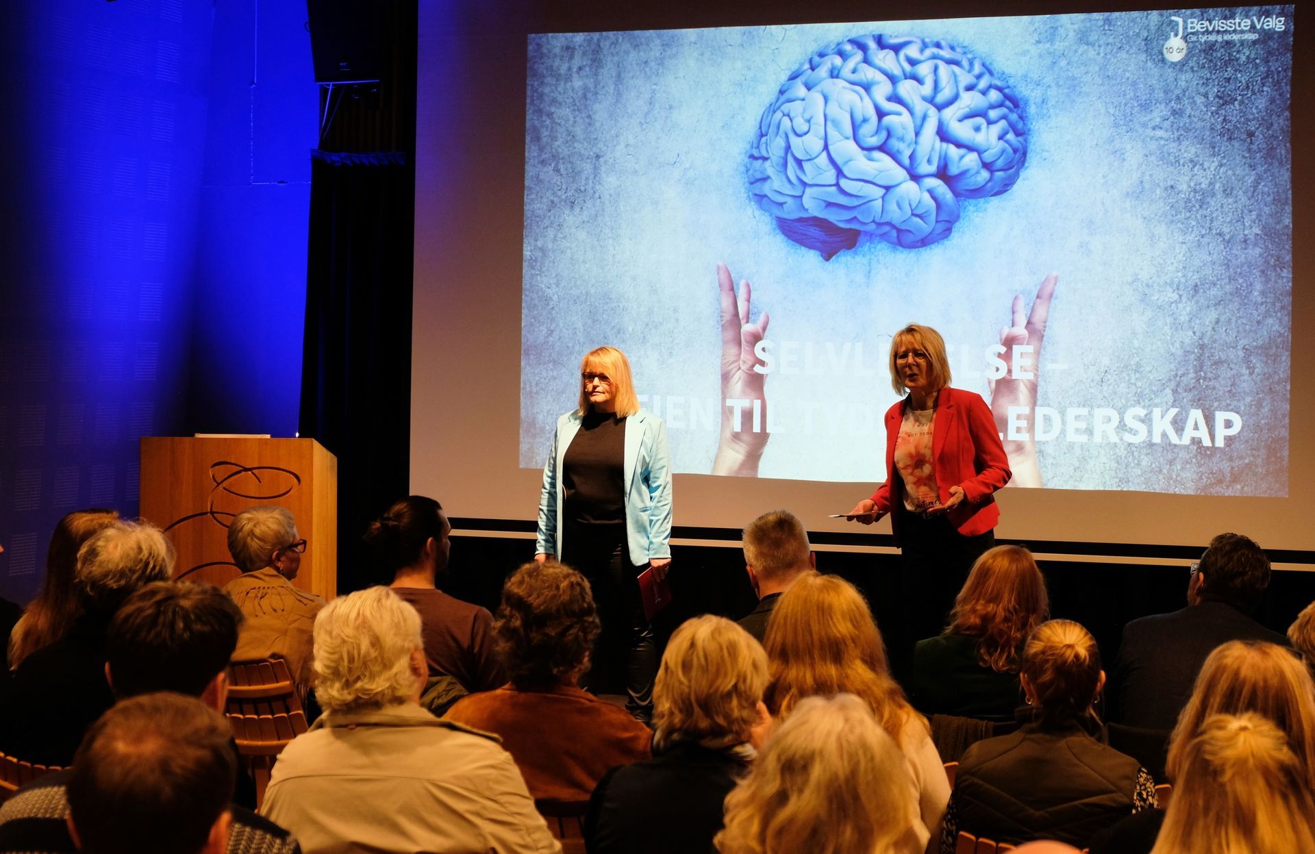 To presentatører står på en scene foran et publikum, med en stor skjerm som viser en hjernegrafikk og ordet «LEDERSKAP».