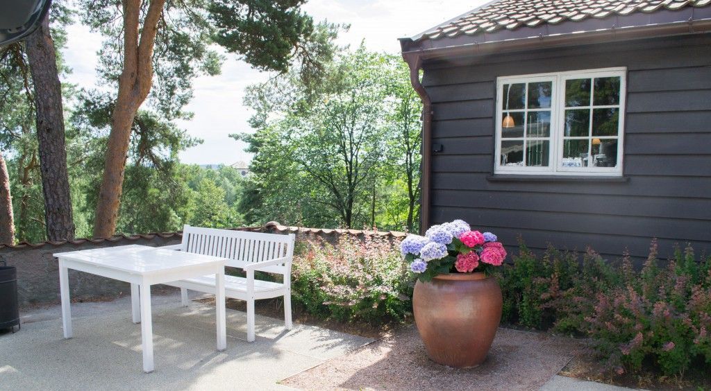 En uteplass med en hvit benk og et bord ved siden av en mørk trehytte, med en potteplante med rosa og blå blomster.