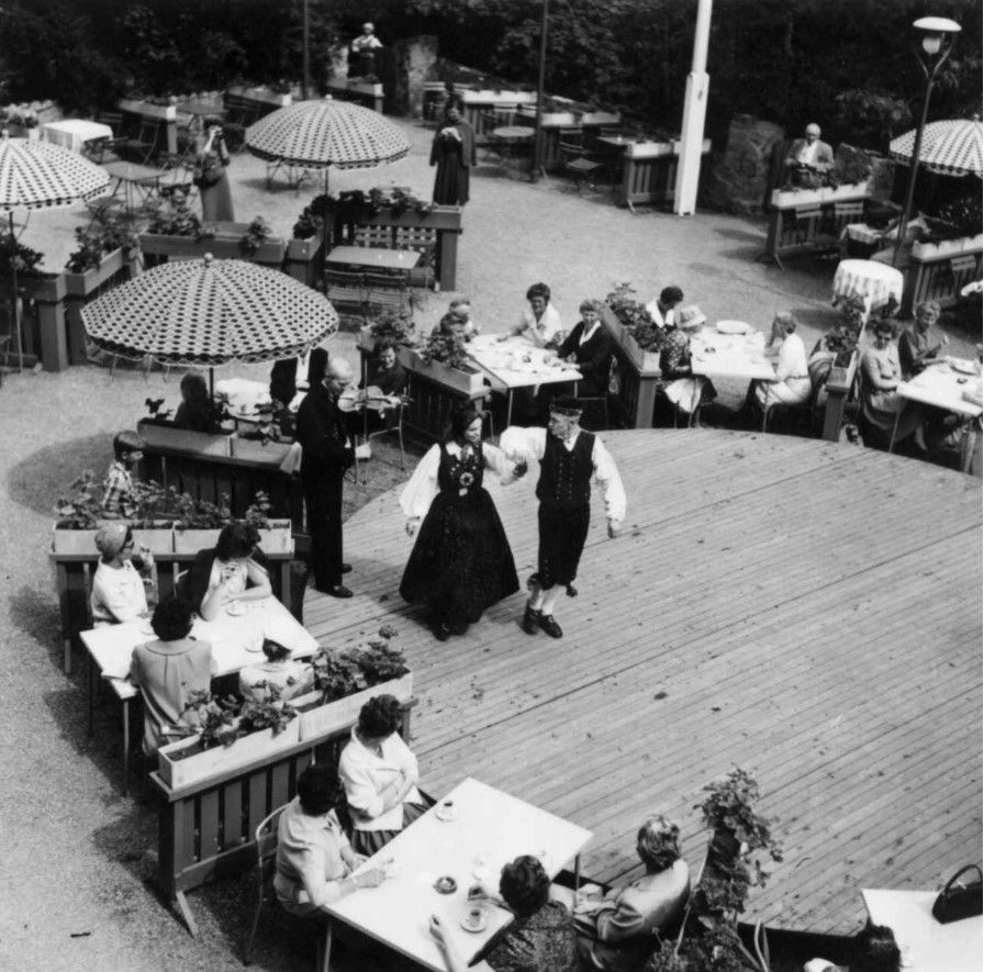 Et par i tradisjonelle folkedrakter danser på en trescene på en restaurantterrasse mens gjestene spiser ved bordene. Gjestestuene er en historisk opplevelse. 
