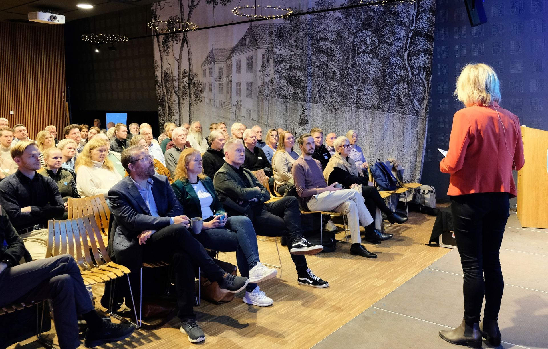 Taler henvender seg til sittende publikum i et konferanserom med en veggmaleri i bakgrunnen