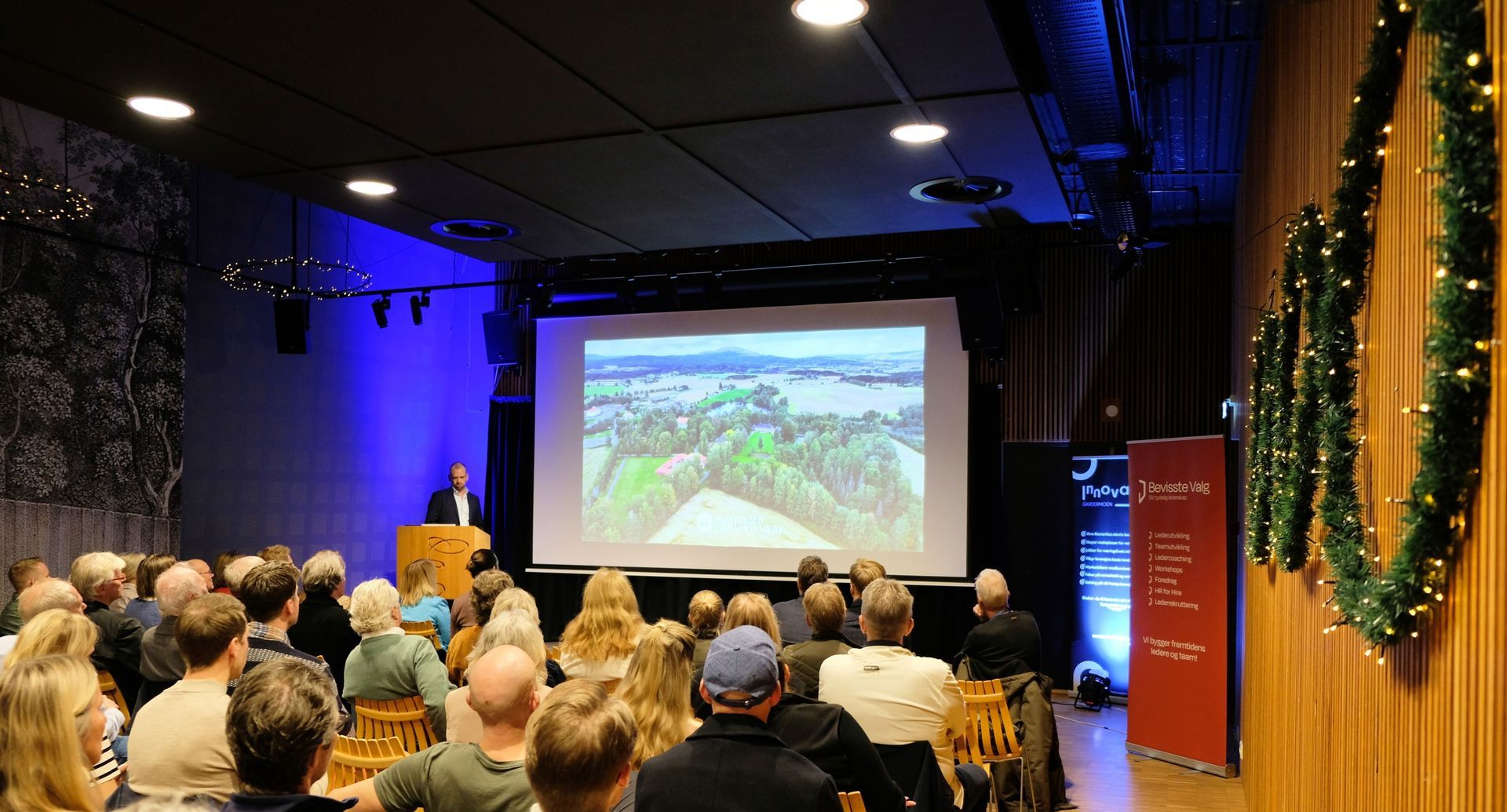 Folk i et konferanserom lytter og smiler. Noen har på seg forretningsantrekk.
