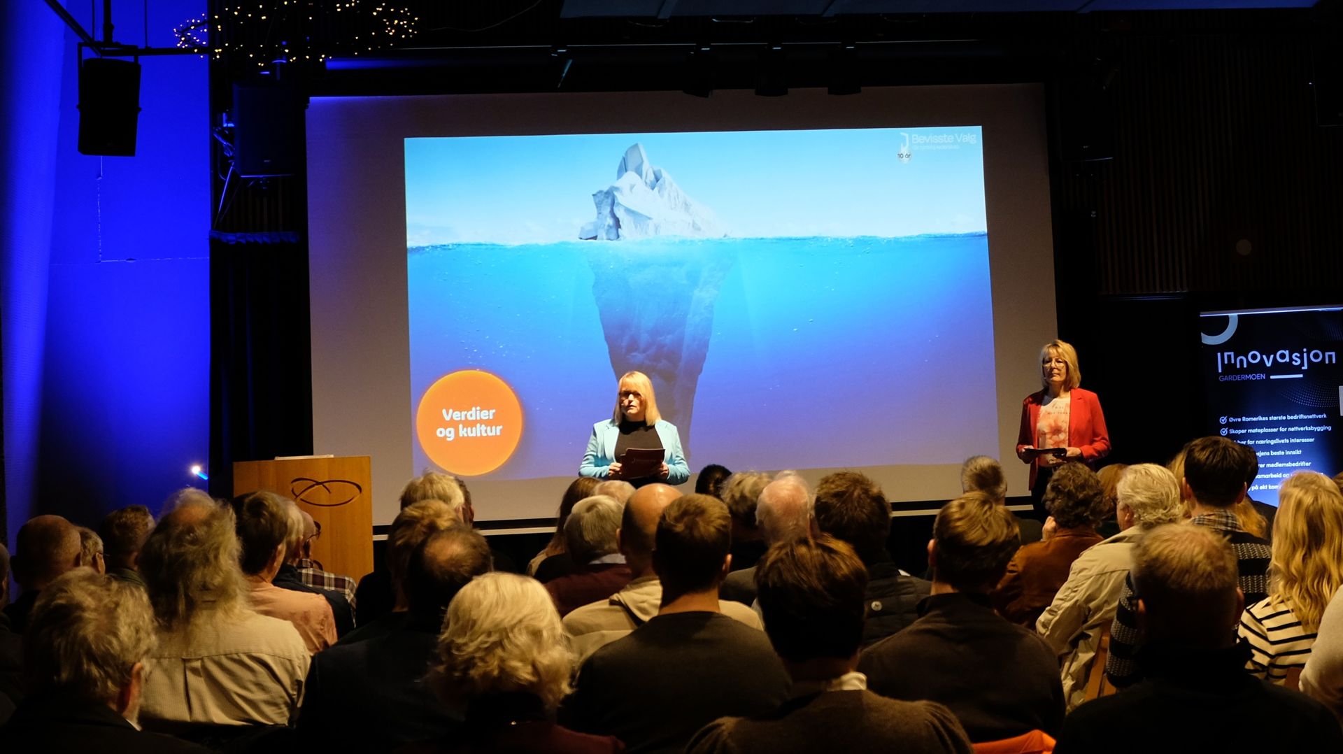 En programleder står på en scene foran et publikum, med en stor skjerm som viser et isfjellgrafikk bak seg.