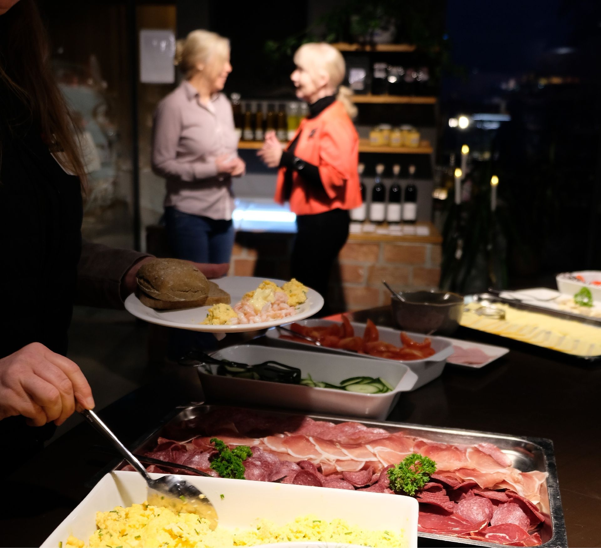 En person serverer frokostretter fra en buffékø mens to andre samtaler i bakgrunnen av et dunkelt, varmt rom.