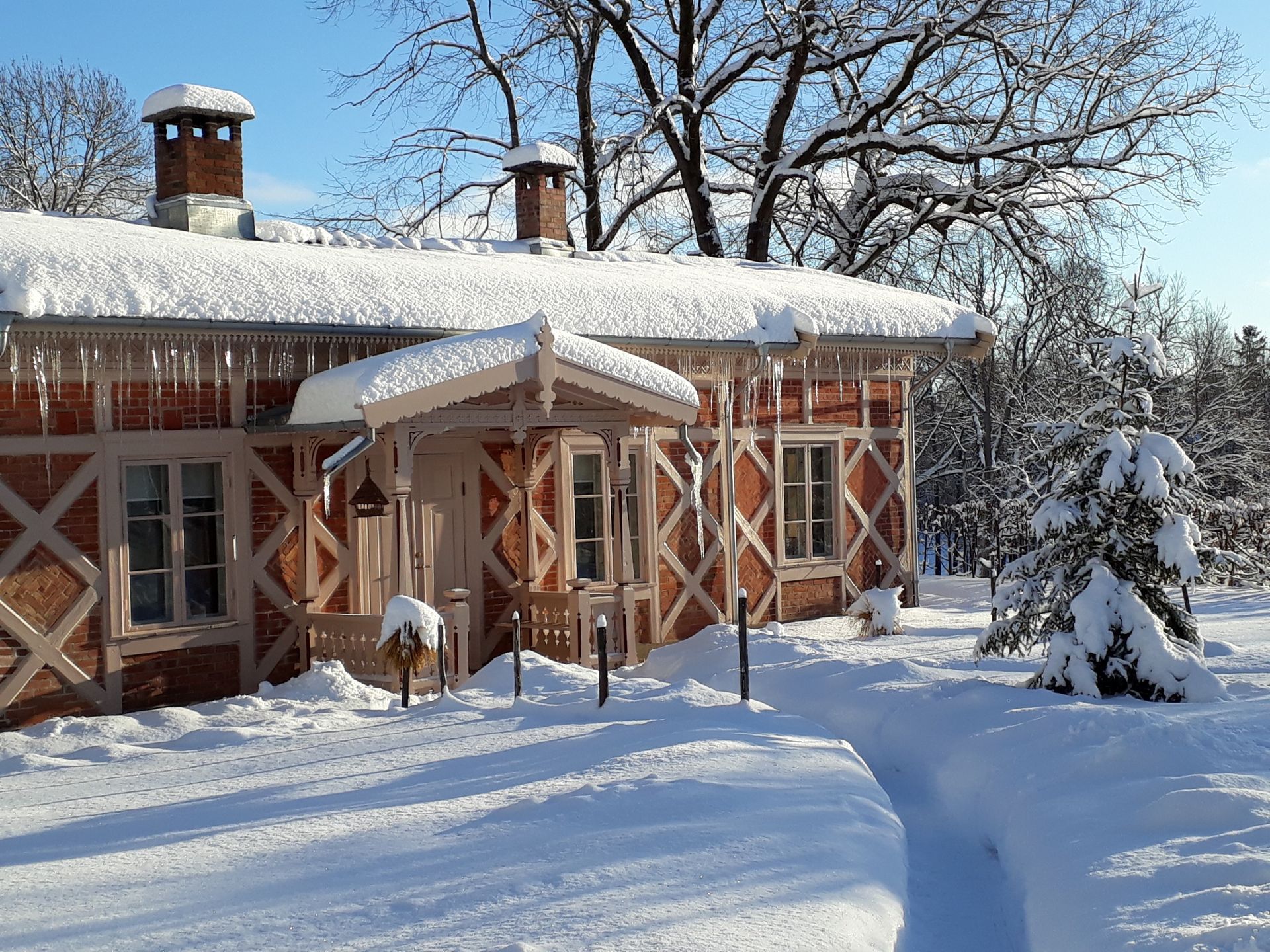 En hytte i rød murstein med hvit bindingsverk og et snødekt tak ligger i et snødekt landskap under en klar blå himmel.