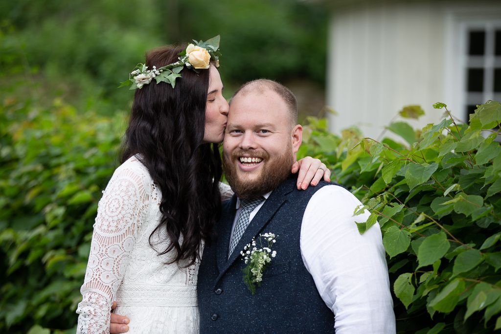 Par poserer utendørs, brud kysser brudgommen på kinnet i en hage.