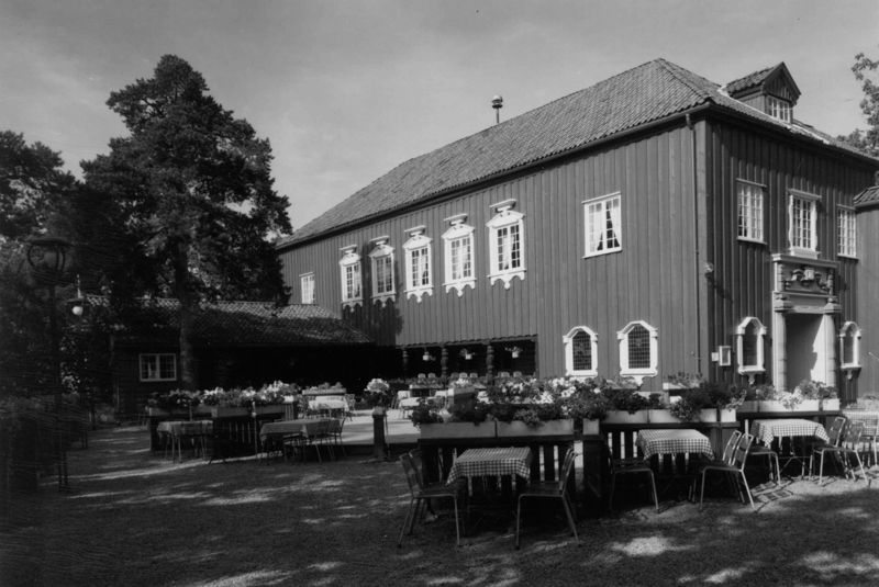 En toetasjes trebygning med en utendørs restaurantterrasse med bord og stoler under trær.
