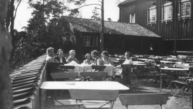 En gruppe mennesker sitter ved bord på en utendørsterrasse foran en rustikk trebygning med hvite vindusrammer.