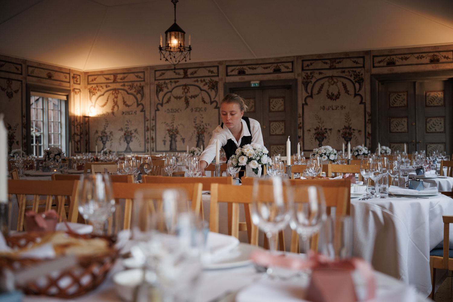 En servitør dekker bordet i en spisesal med blomstermalerier og rader med hvitkledde bord klargjort til et arrangement.