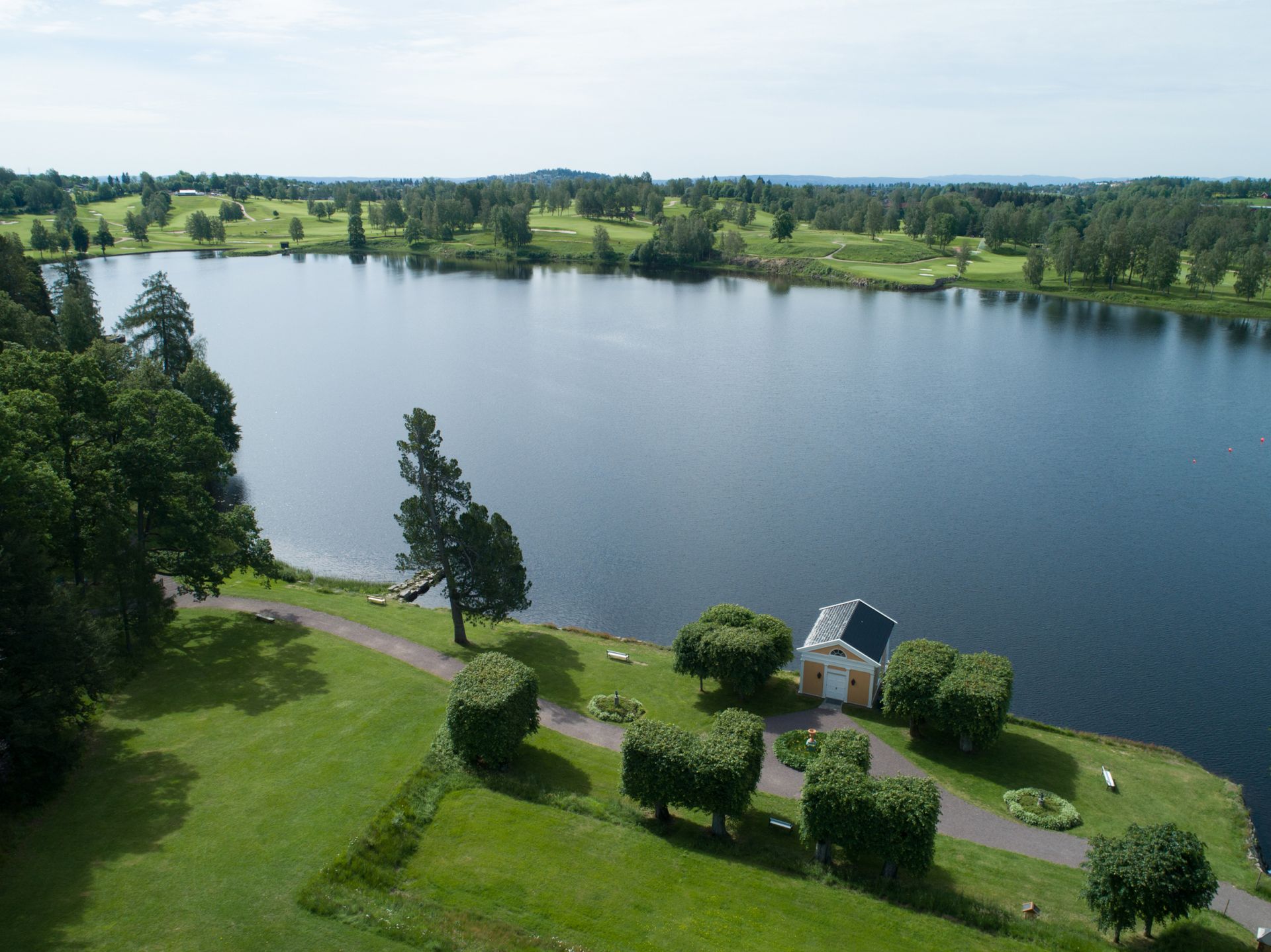 En innsjø omkranset av grønne trær og en plen med pent trimmede hekker og en liten hvit bygning langs bredden.