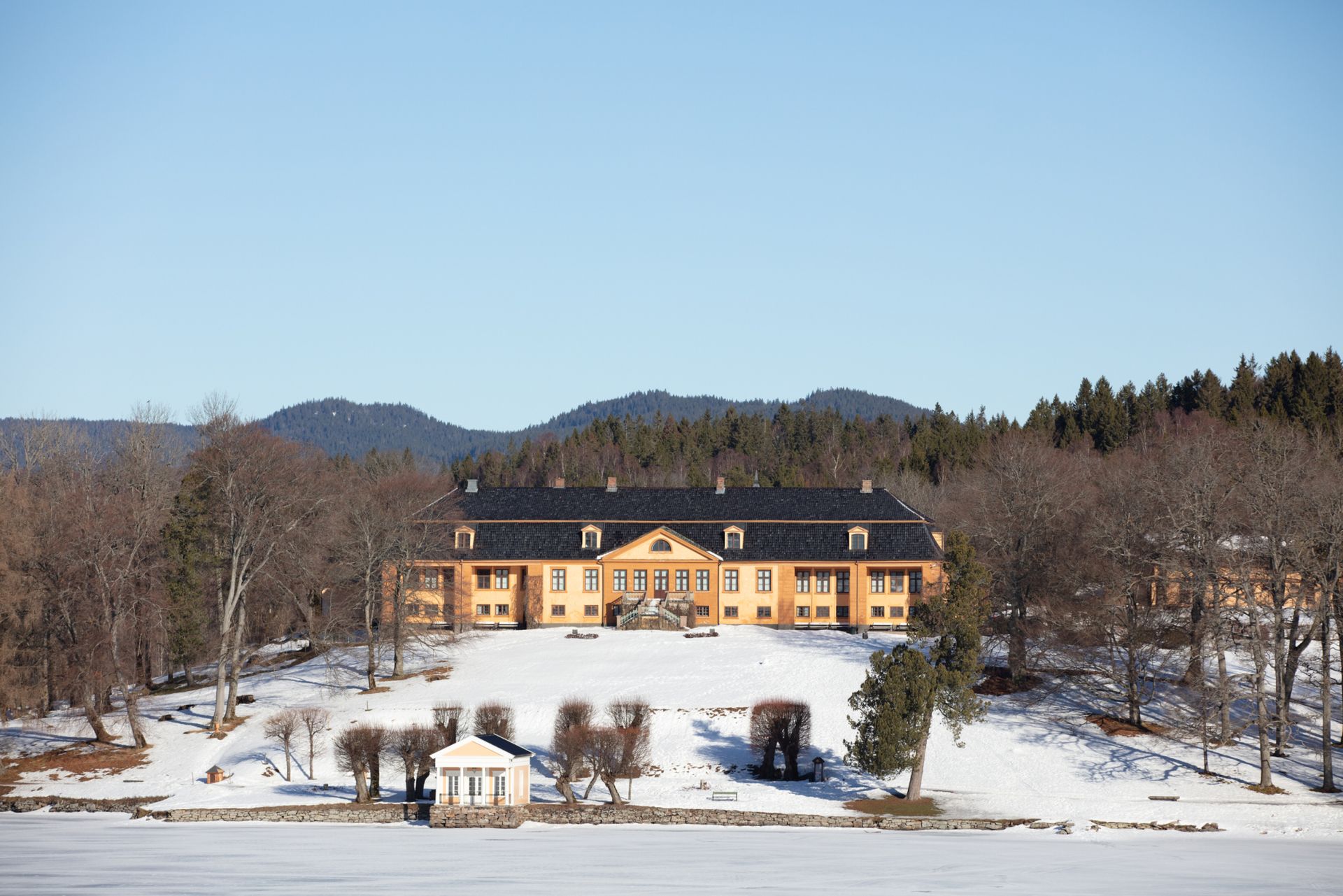 Et stort, gult herregård ligger på en snødekt ås over en frossen innsjø, innrammet av nakne trær og fjerne skogkledde fjell.