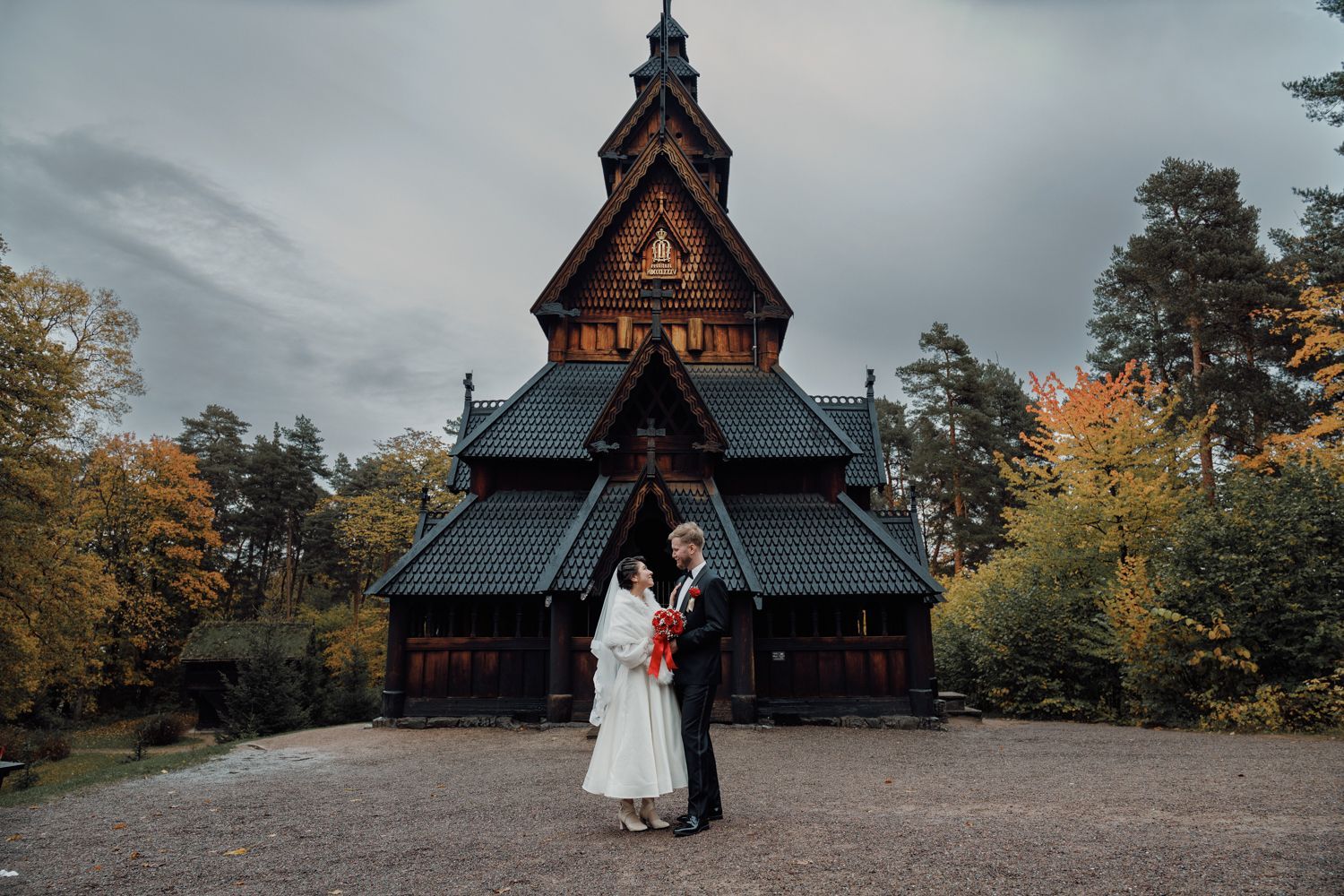 Et brudepar som står foran en historisk stavkirke av tre omgitt av høsttrær. 