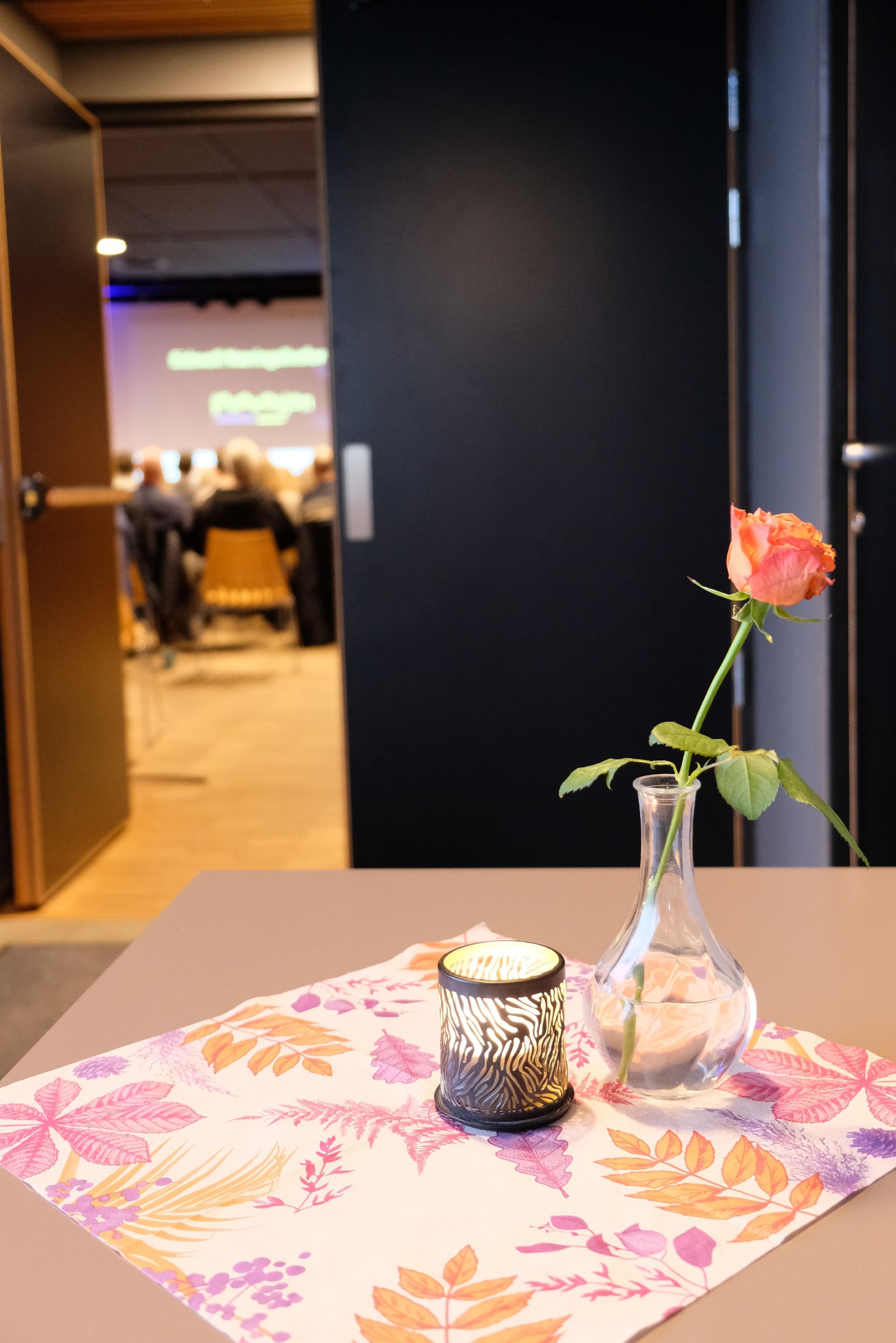 En rosa rose i en glassvase og en liten lykt står på et blomstermønstret bord foran et uskarpt konferanserom.