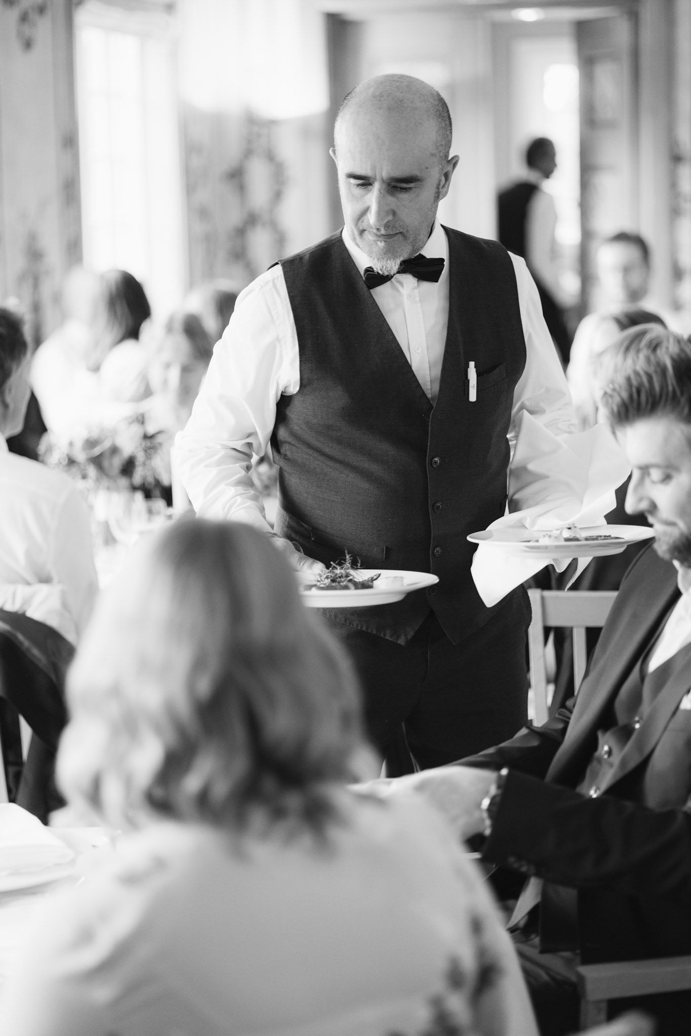 En servitør i vest og sløyfe serverer en tallerken til en spisende person ved et bord i en formell, godt opplyst restaurant.