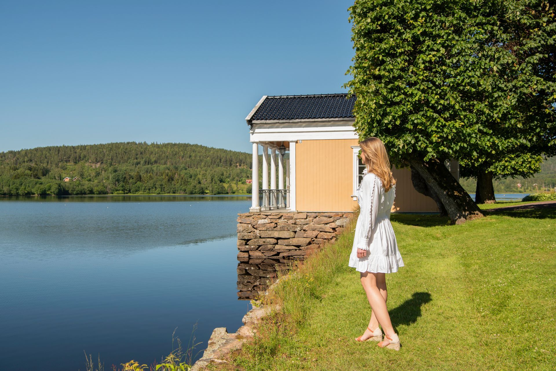 Kvinne i en hvit kjole som står ved en rolig innsjø ved siden av en steinmur og et velstelt tre
