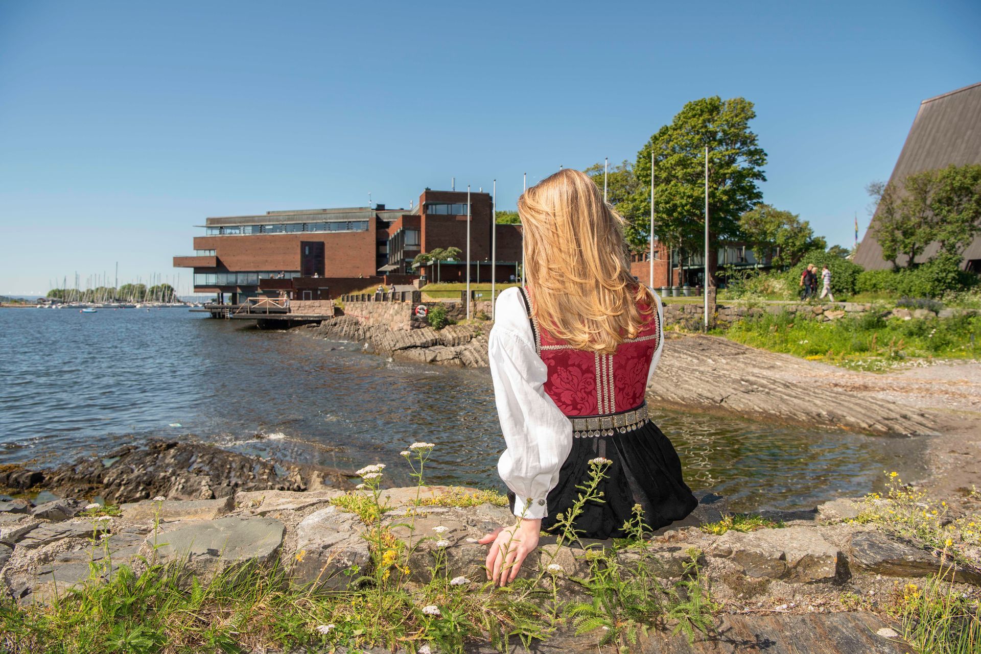 En person i en tradisjonell rød og svart folkedrakt sitter på en steinete strand med utsikt over en innsjø og en moderne murbygning.