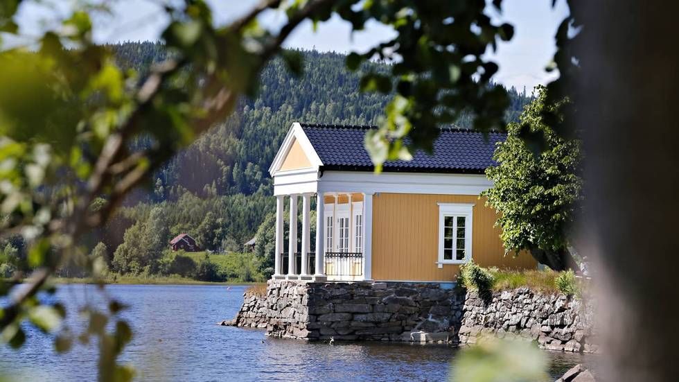 Gul hytte ved innsjøen med hvit veranda og steinfundament, innrammet av trær på rolig vann
