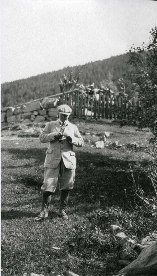 En person i tweeddress og sixpack står i et gresskledd område med en steinmur og en skogkledd åsside i bakgrunnen.