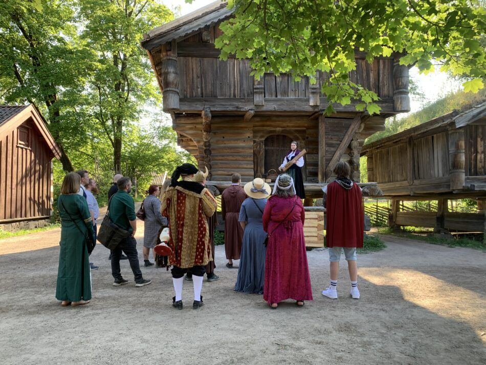 En gruppe mennesker i historiske klær samles på en gresskledd gårdsplass for å se en utøver på en rustikk trekonstruksjon.