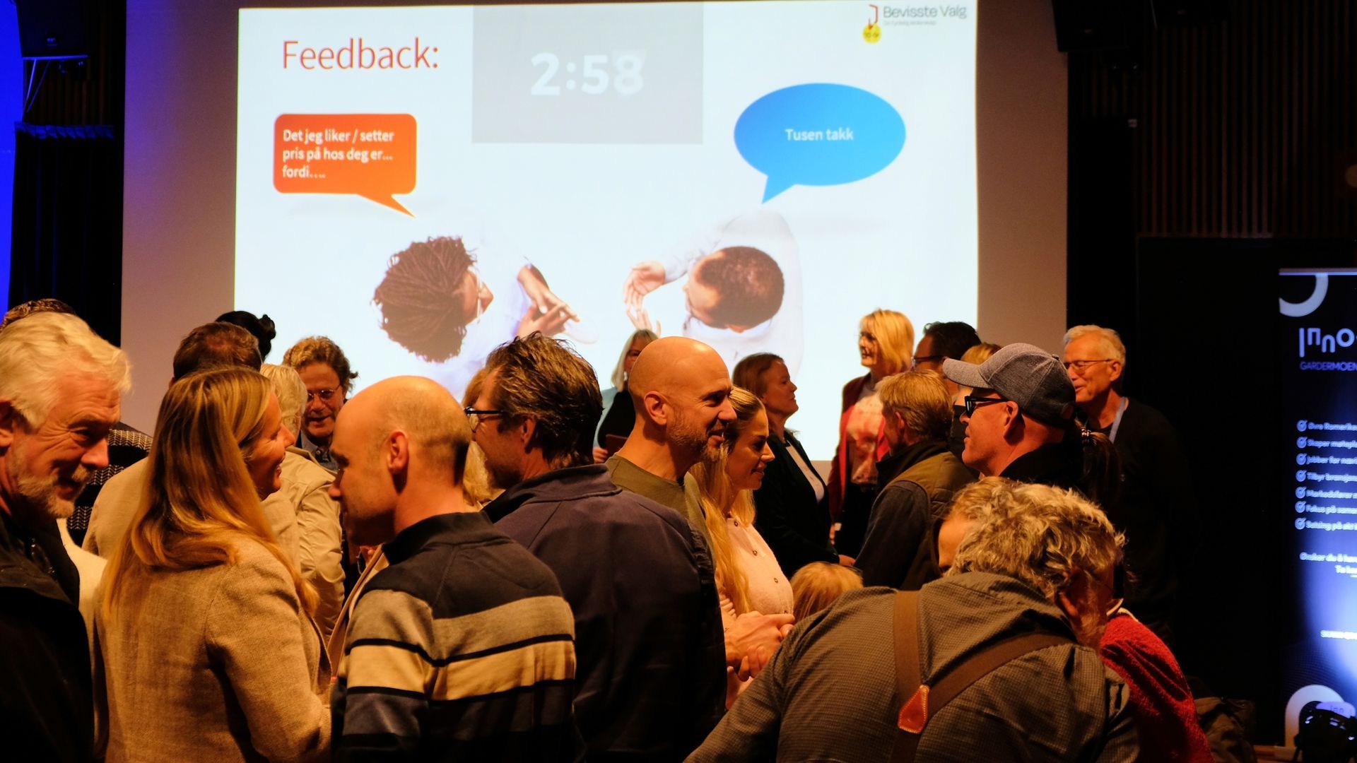 En gruppe mennesker som nettverker i et svakt opplyst rom foran en presentasjonsskjerm om å gi og motta tilbakemeldinger.