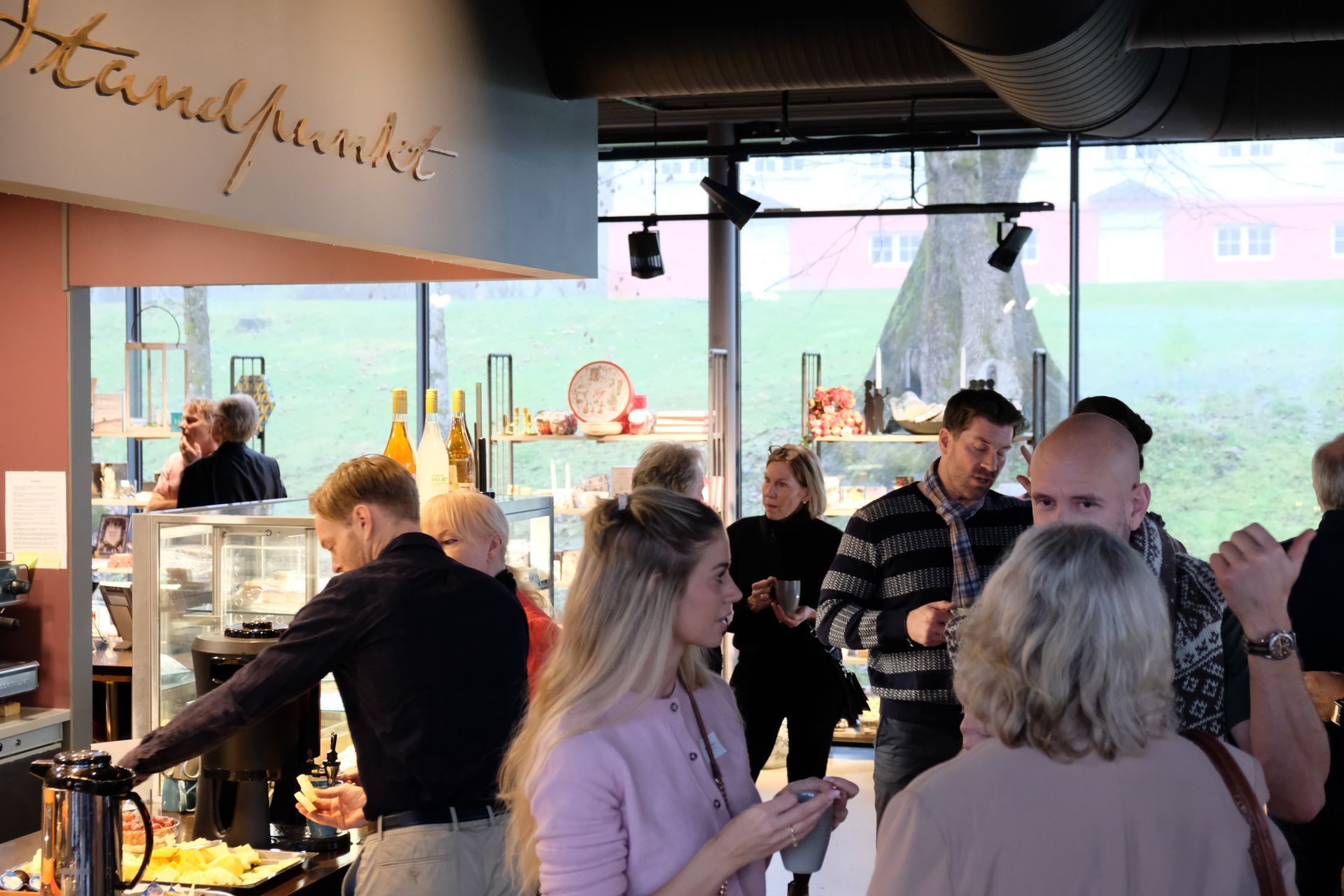 Folk prater og drikker i en lys kafébar med store vinduer og hengende lamper.