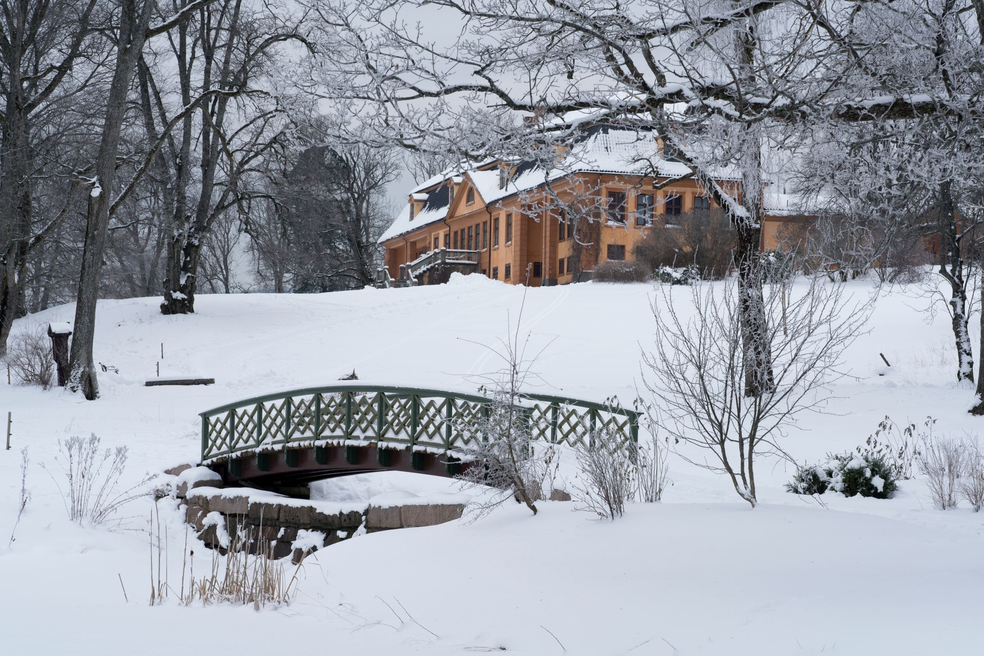 Snødekt park med en grønn bro og et oransje hus blant bladløse trær