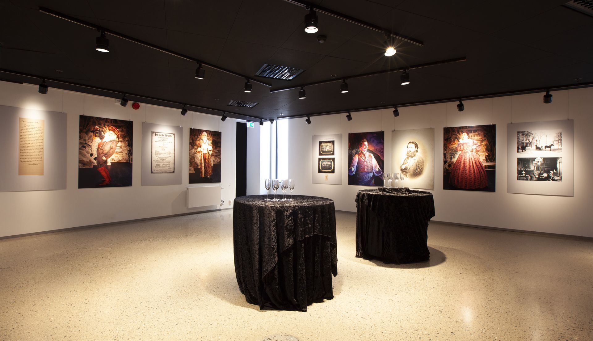 Et kunstgalleri med hvite vegger som viser innrammede bilder og to runde bord med svarte duker på et steingulv.