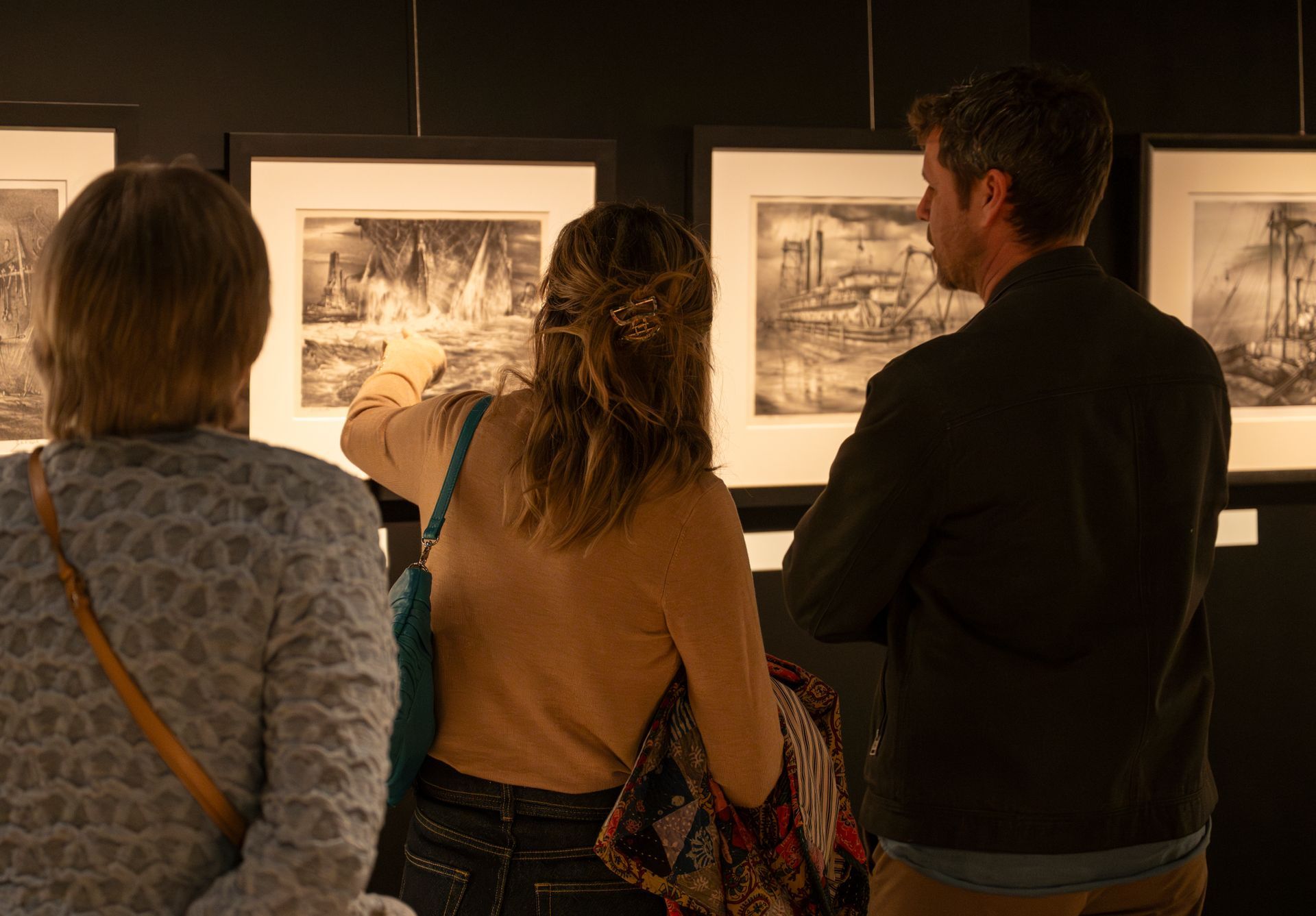 Tre personer ser på innrammede svart-hvitt-tegninger i et kunstgalleri, mens én person peker mot kunstverket.