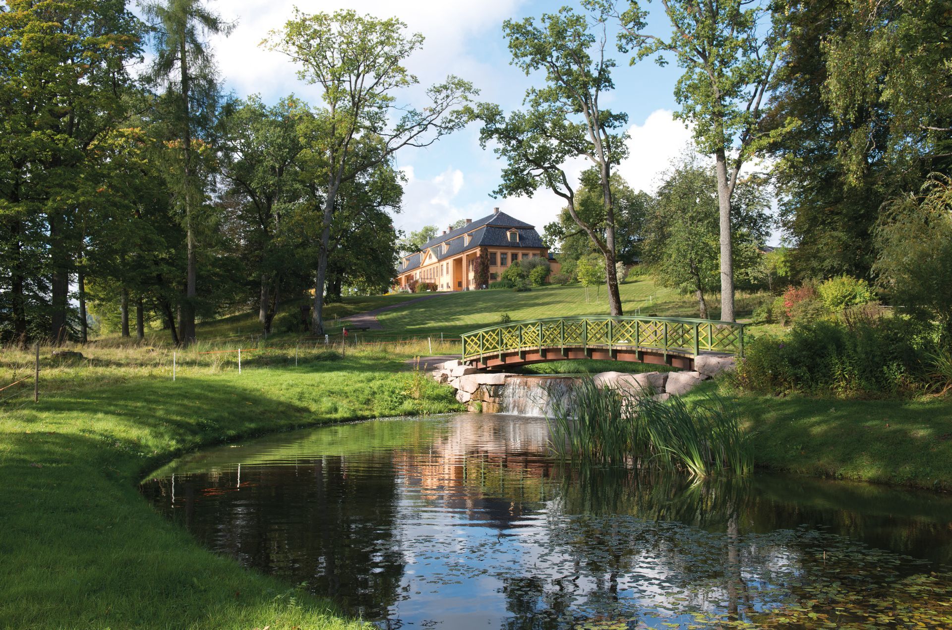 Solrik park med en liten bro over et tjern og Bogstad Gård blant trær i bakgrunnen