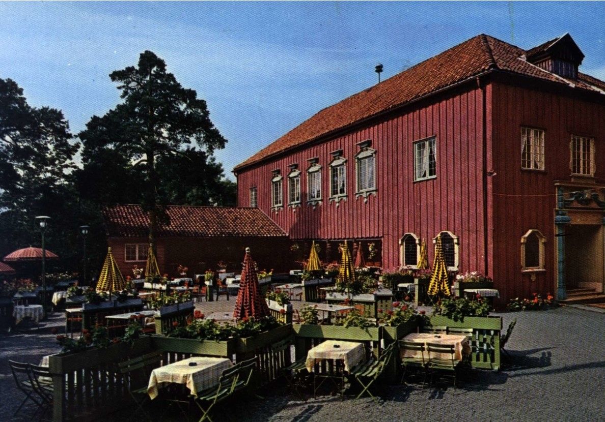 Utendørs spiseplass med bord og gule parasoller foran en stor, toetasjes rød trebygning under en blå himmel.