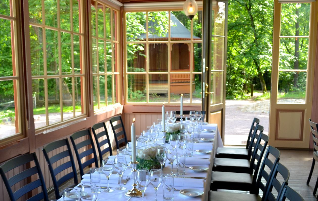 Et langt spisebord dekket til et arrangement i en solfylt hagestue med glassvegger, med utsikt over grøntområder og et lysthus.