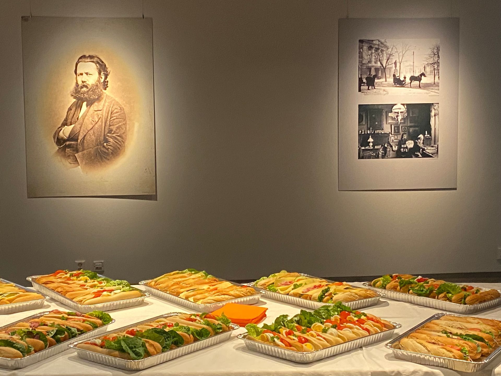 Brett med smørbrød arrangert på et bord i et galleri med to historiske portretter og fotografier på veggen bak.