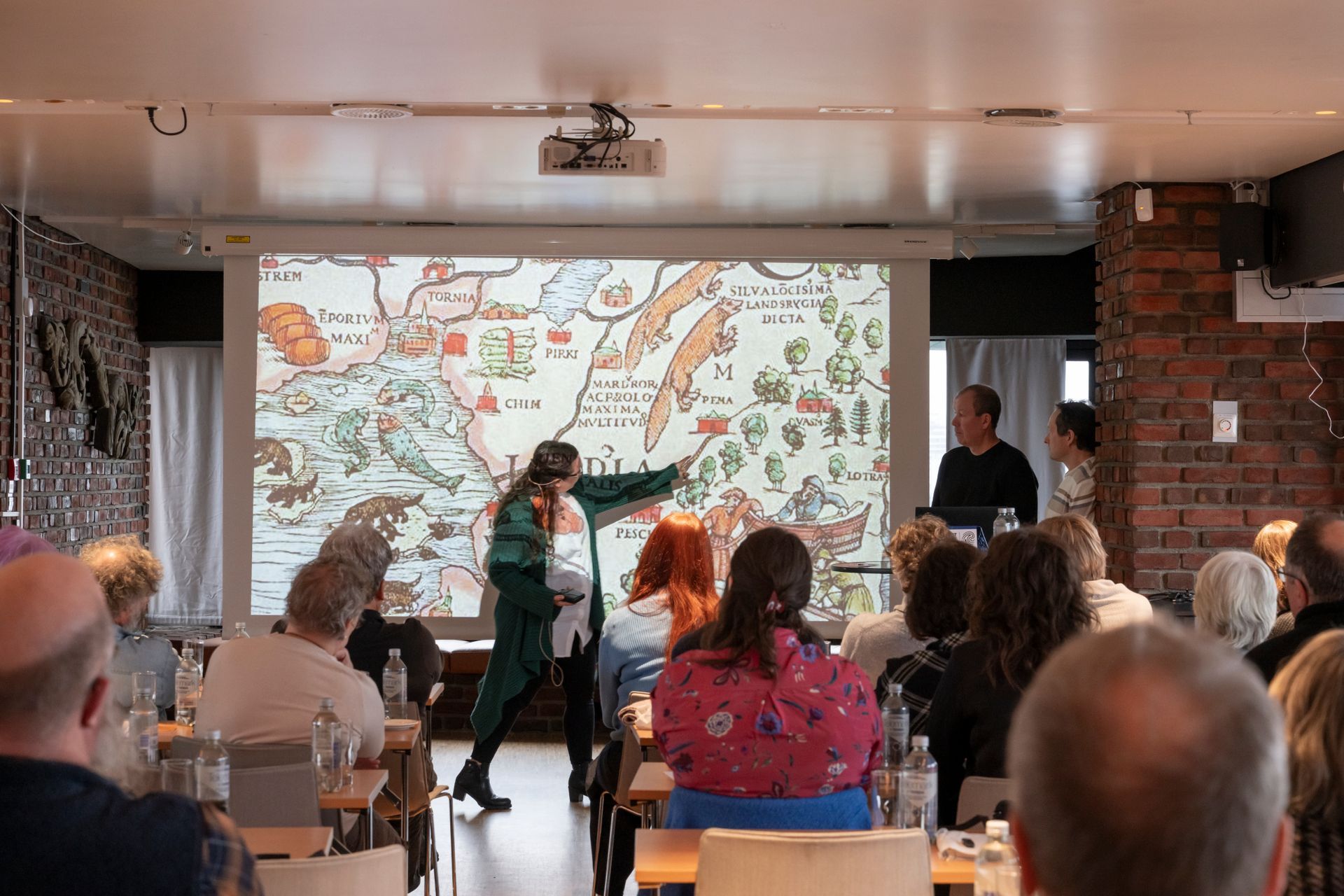 En presentatør står foran et projisert historisk kart og peker på en detalj for et publikum i et rom med murvegger.