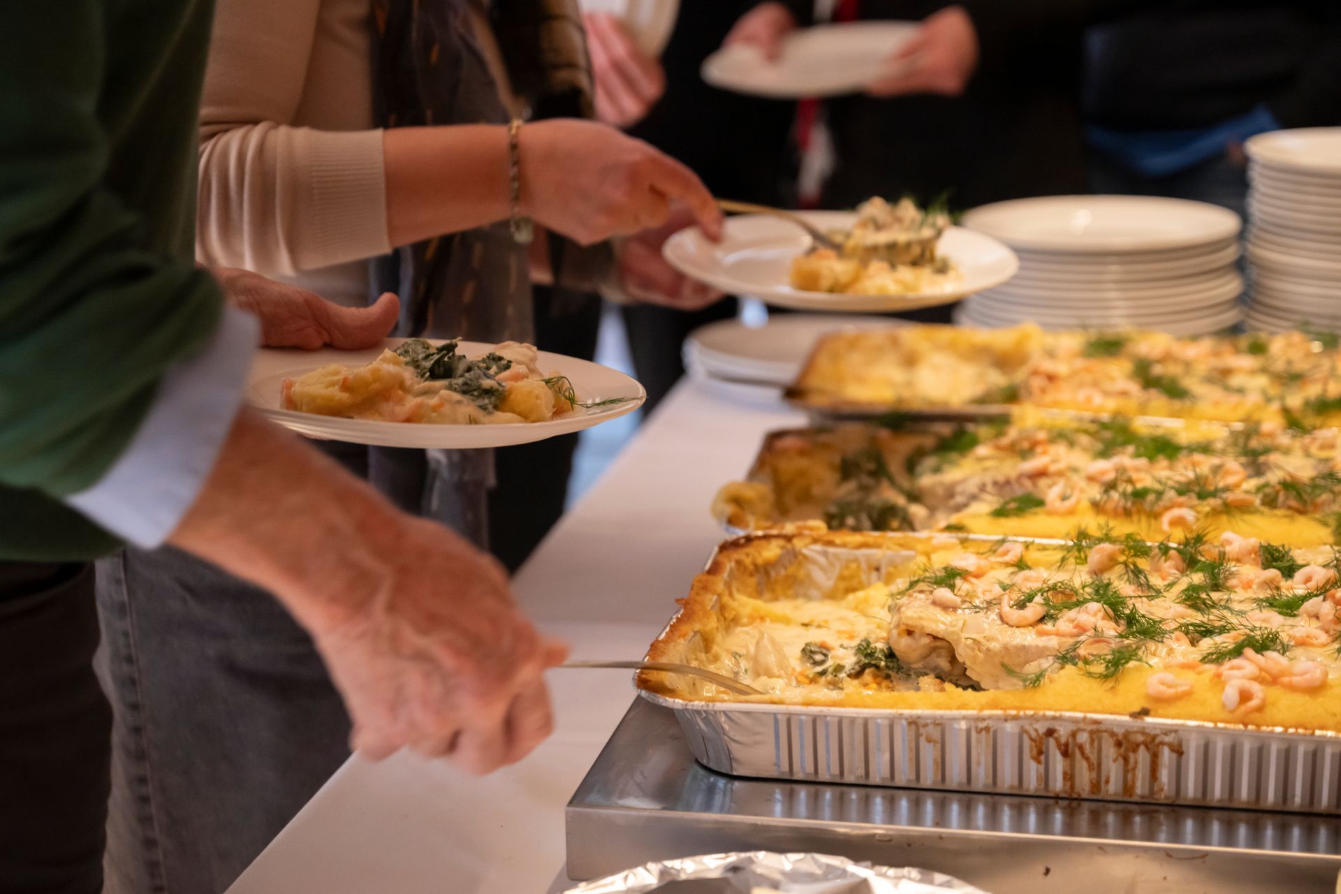 Folk som serverer seg selv fra en buffé med store metallbrett fylt med mat på et cateringarrangement.