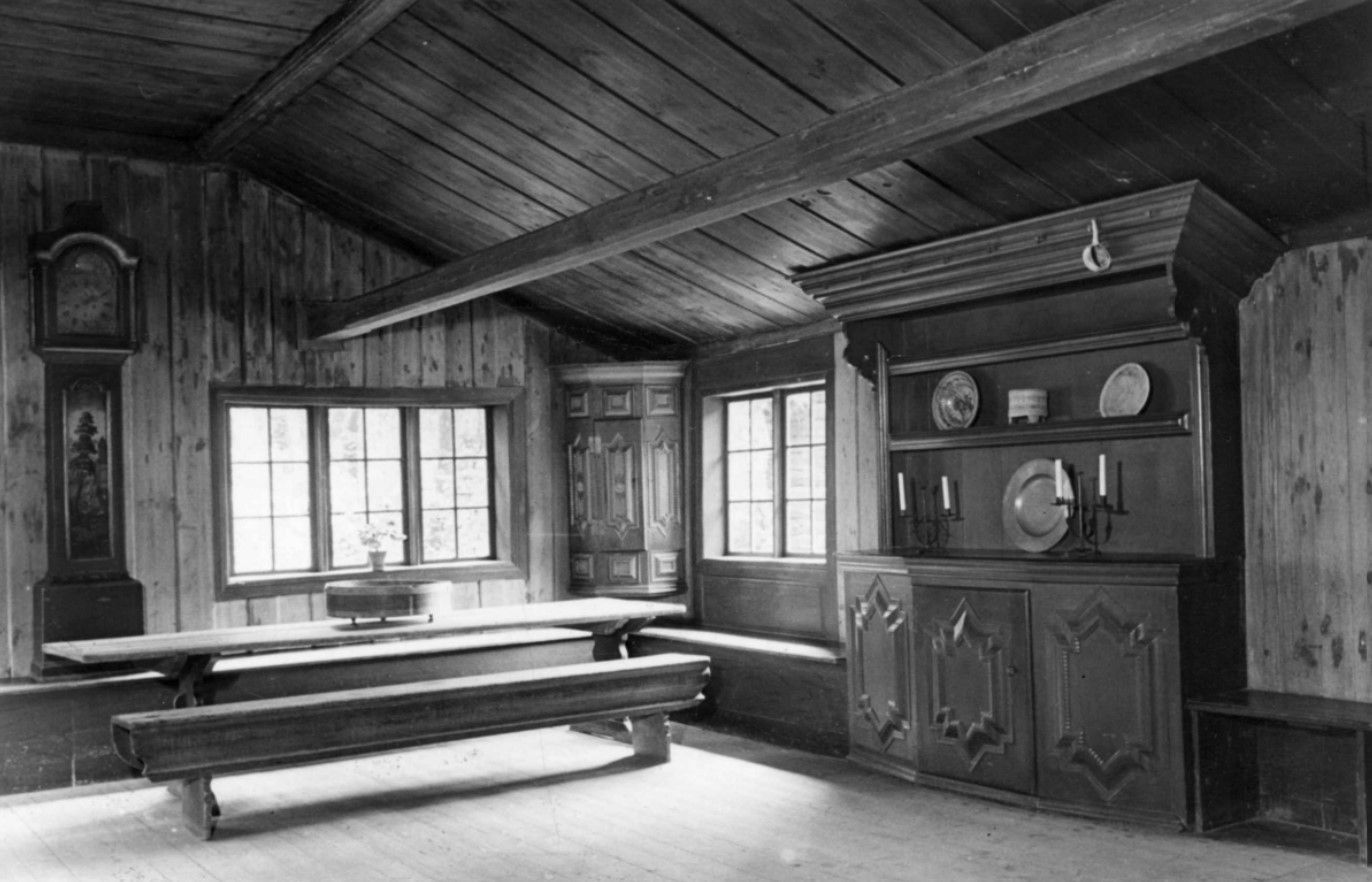 Et rustikt rom med trepanelvegger, et langt trebord med benker, en høy klokke og et utsmykket bur med tallerkener.