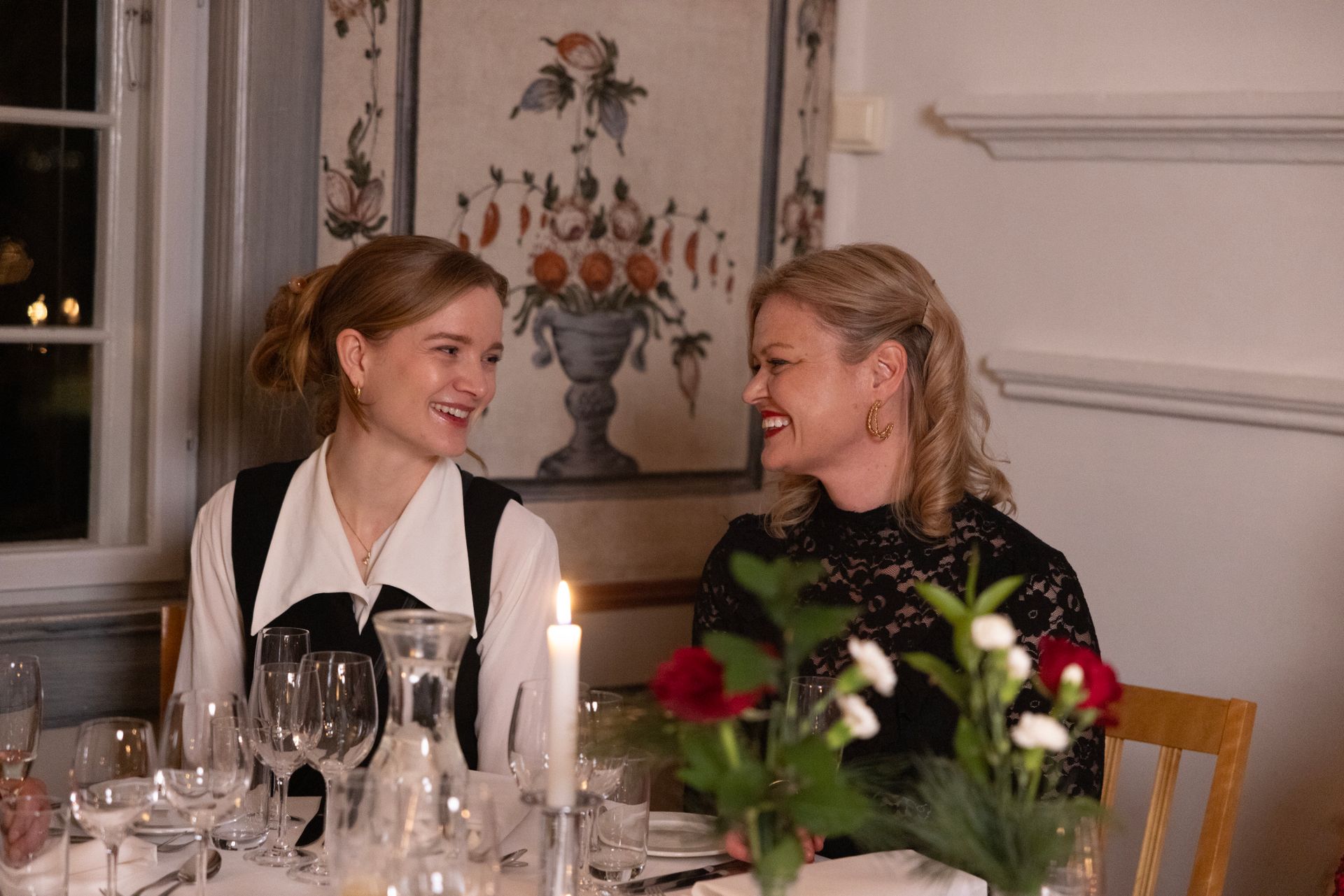 To personer sitter ved et bord med levende lys, smiler og ser på hverandre i et varmt opplyst rom med blomsterveggkunst.