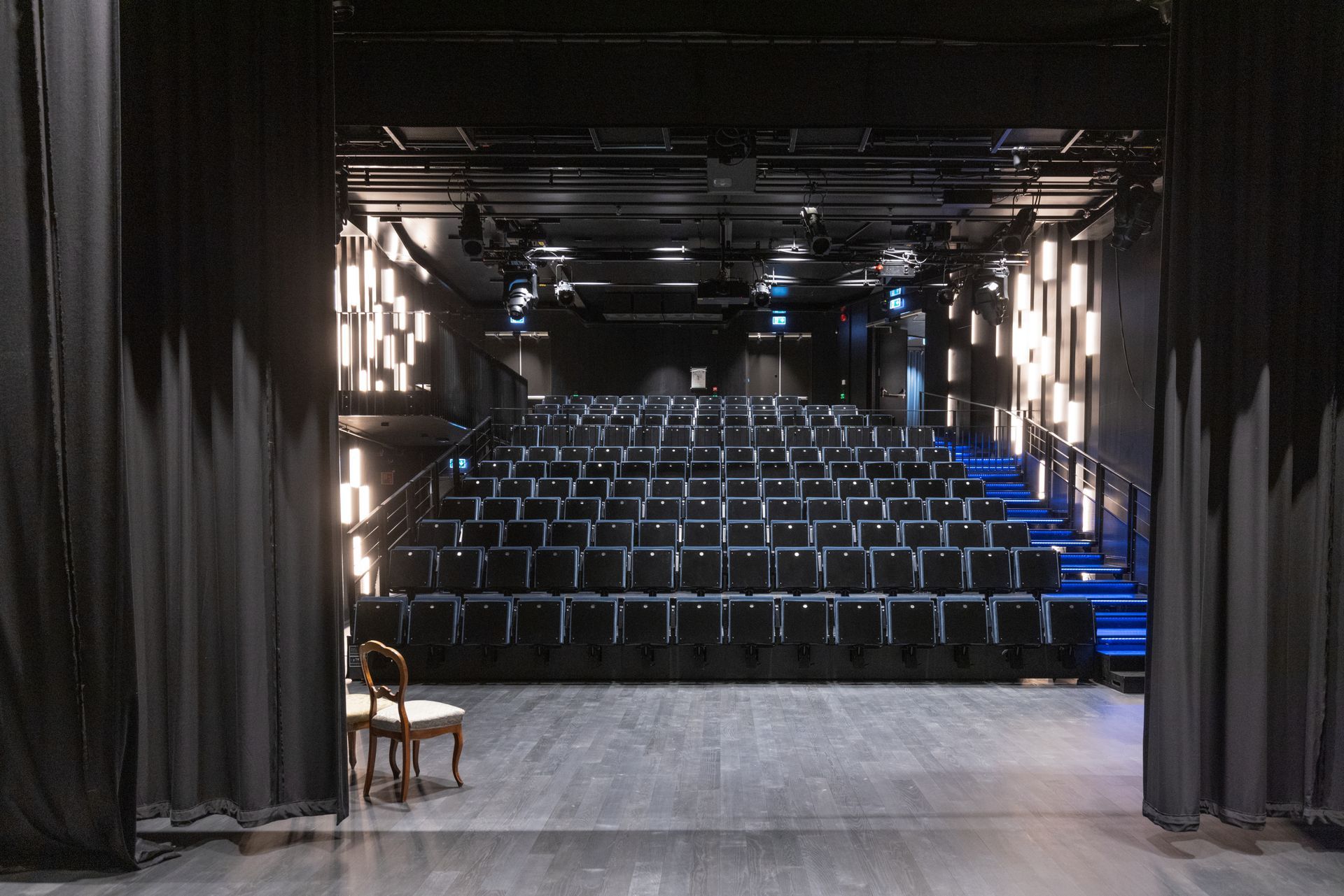 Utsikt fra en scene med utsikt over et mørkt, moderne teater med rader med svarte seter, opplyst av vertikale hvite vegglamper.