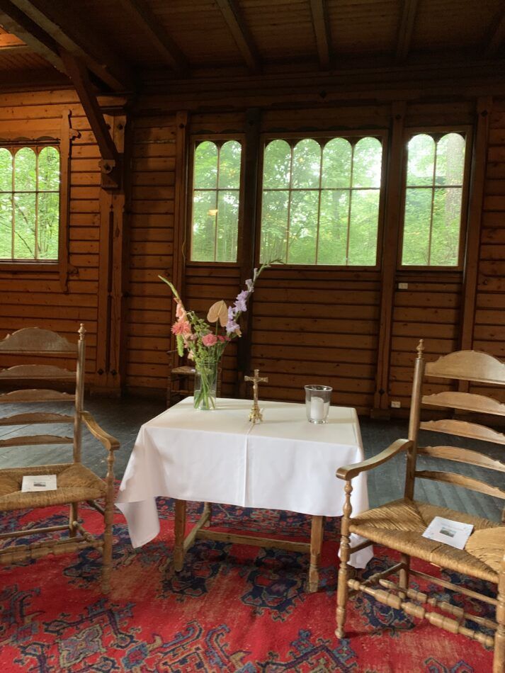 To trestoler flankerer et lite bord med en hvit duk, et blomsterarrangement, et kors og et stearinlys i et rustikt rom.