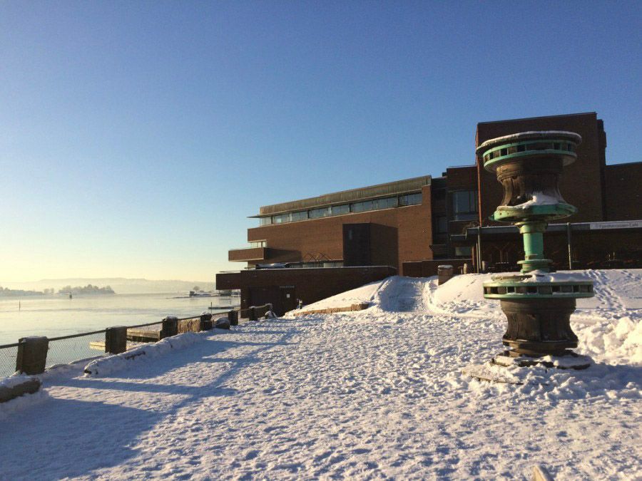 Snødekt havnefront med en etasjevis skulptur i forgrunnen og en moderne murbygning under en klar blå himmel.