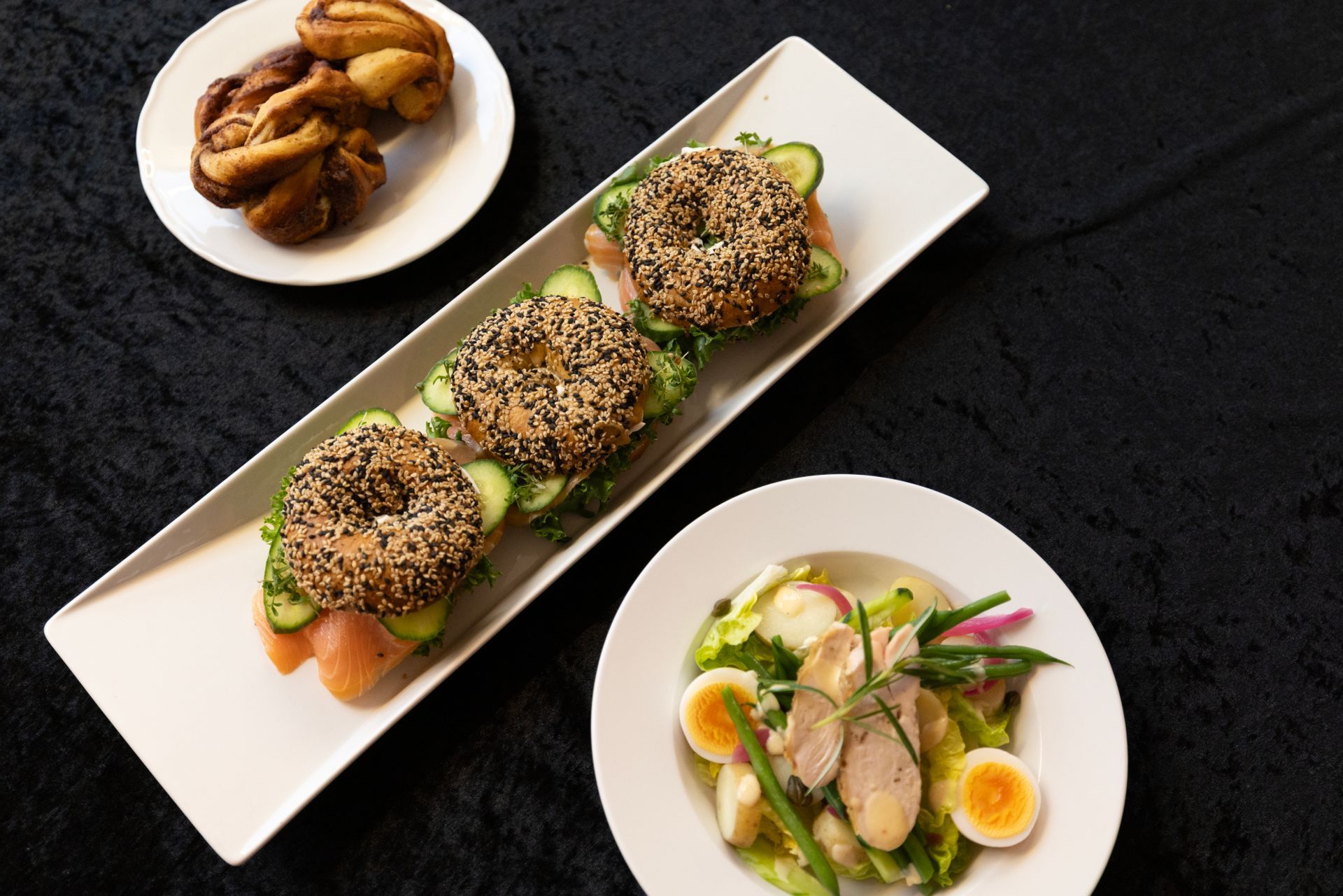 Assorterte gourmetforretter på et mørkt bord, inkludert sesamdekkede sandwicher, stekte boller og en salat.
