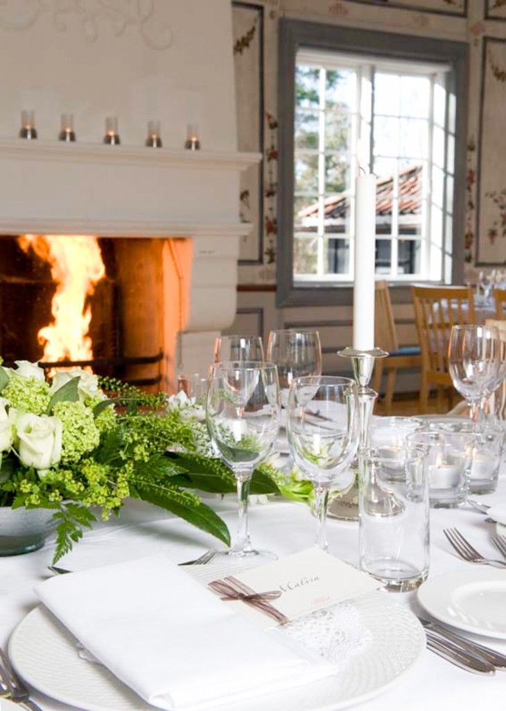 Elegant spisebord dekket ved siden av en tent peis i et lyst rom med hvite blomster