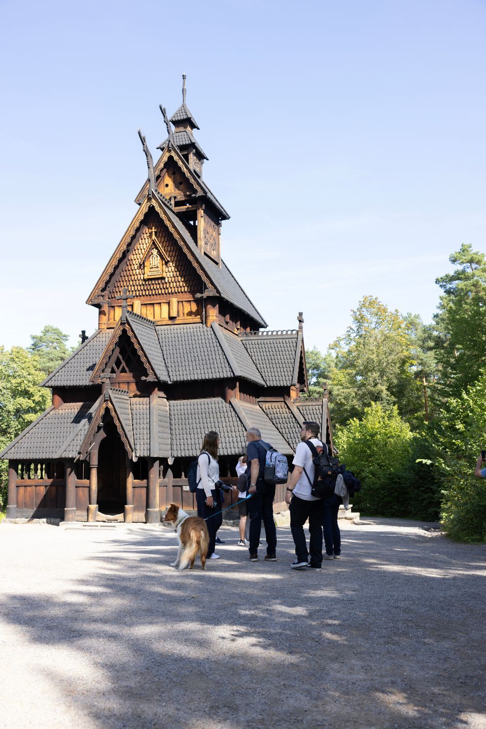 Stavkirke i tre, utsmykkede utskjæringer, folk som går mot inngangen, solrik dag.