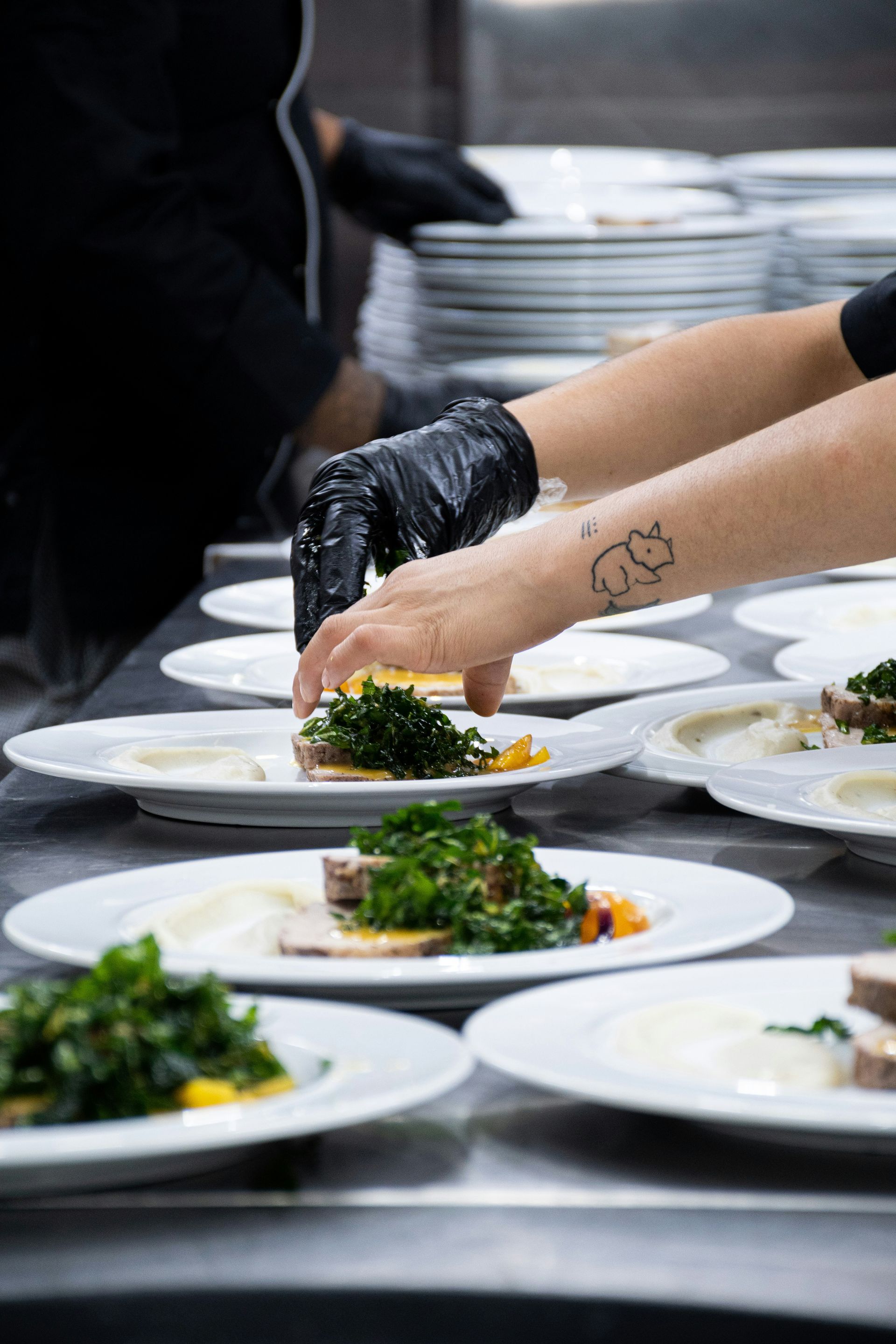 Kokk setter mat opp på hvite tallerkener på et restaurantkjøkken; svarte hansker, stablede tallerkener.