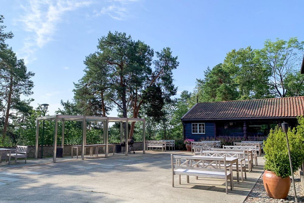 En utendørsterrasse med hvite benker og en pergola i tre, som ligger ved siden av en mørkeblå bygning omgitt av trær.