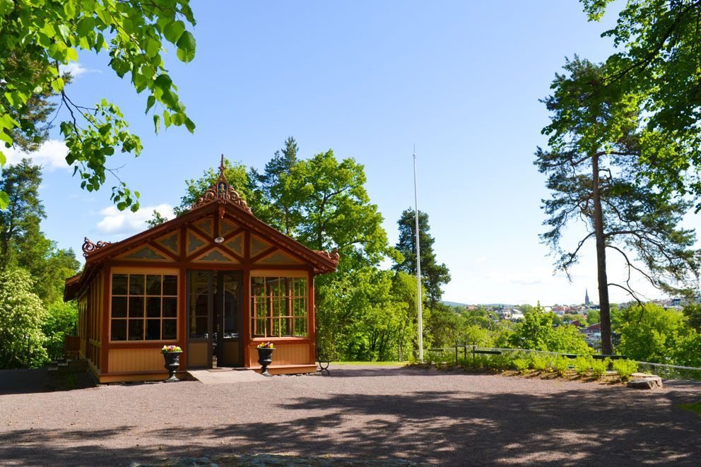 Et lysthus i tre med glassvegger står på en gruslysning i en solrik park med trær og utsikt over byen i det fjerne.