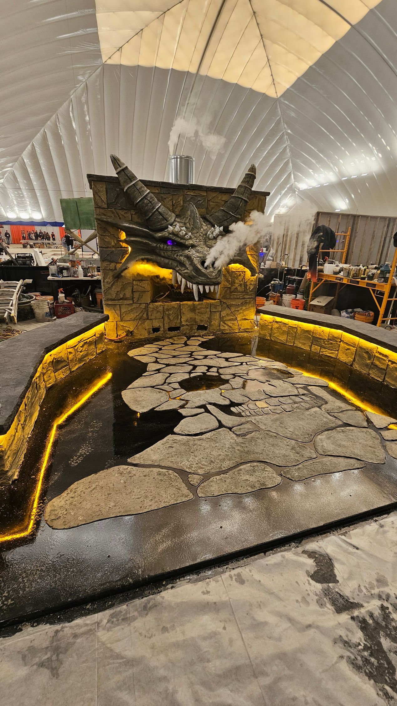A large, glowing dragon sculpture sits above a dark stone pool, with smoke emitting from its mouth in a studio setting.