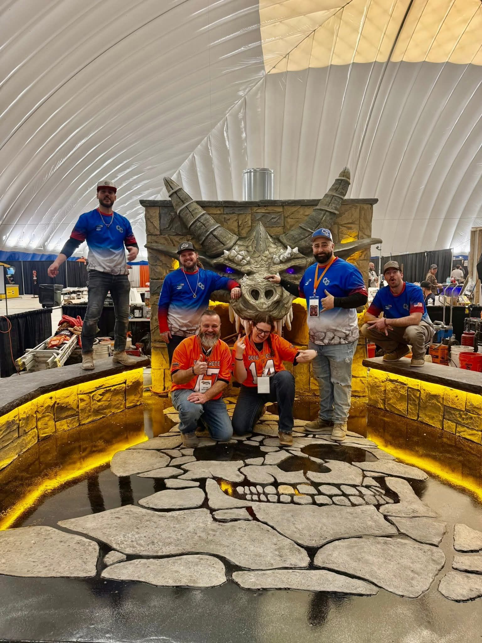 Six people pose in front of a large, stone-style dragon head sculpture inside an exhibition tent.