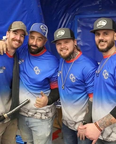 Four people in matching blue and white team shirts and baseball caps smile together in front of a blue backdrop.